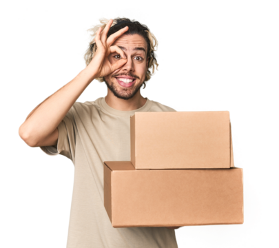Young man with cardboard boxes in studio excited keeping ok gesture on eye.