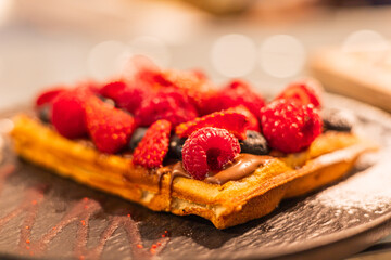 A delicious waffle topped with fresh raspberries and blueberries on a dark plate. The background is softly blurred, enhancing the focus on the dessert.