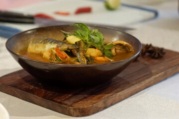 Close up of traditional Indonesian fish soup with herbs, chili, and vegetables, served in a rustic bowl on wooden board