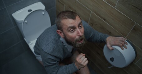 Obraz premium Businessman with Tense Facial Expression Suffering from Diarrhea and Indigestion, Sitting on the Toilet. Man Experiencing Upset Stomach, Digestive Discomfort in Office Restroom During Work. High Angle