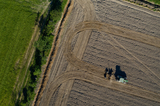 Aerial view of a tractor tilling the rich, dark earth of rice fields, contrasted by the vibrant green of adjacent fields, Vespolate, Piedmont, Italy.