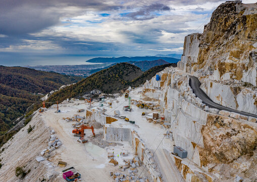 Aerial view of marble quarries reveal stark white cuts against rugged terrain, under a sky streaked with dramatic clouds, Carrara, Tuscany, Italy.
