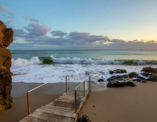 Coastal escape with a rustic wooden staircase descending to the sandy shore as waves crash against the rocks at dusk
