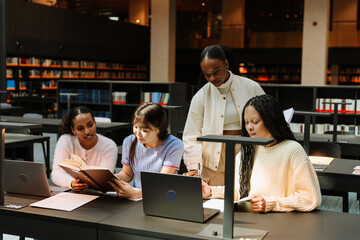 A group of three female students are sitting at a table reading a book and a notebook while a female student stands next to them