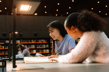 Female student looking at laptop and smiling while sitting at table near female student holding a...