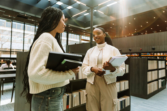 Two female students standing near bookshelves and talking while holding notebooks and a laptop