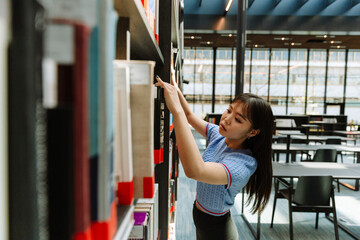 A female student is standing and reading the cover while taking out a book from a bookshelf