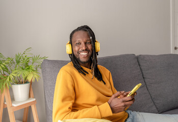 African american man with dreadlocks wearing yellow headphones sitting on sofa using smartphone