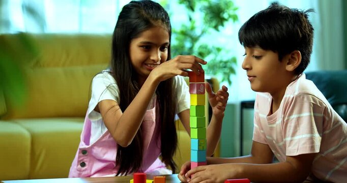 Indian small brother sister building wooden toy tower together at home indoors, Asian young kids siblings enjoying fun childhood activity, spending quality time with family bonding in modern home