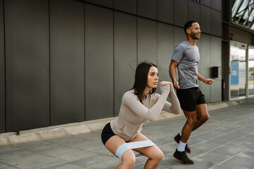 A female athlete squats with a rubber band on her hips while a male athlete jumps rope next to her