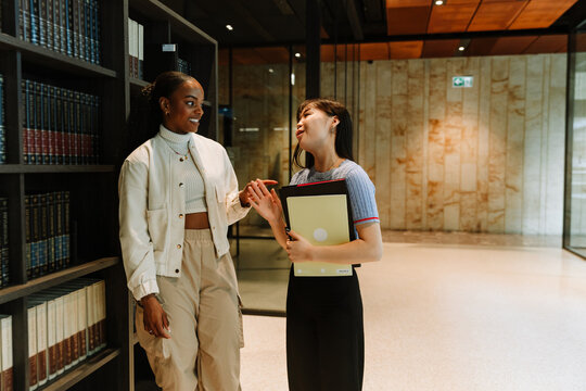 Two female students are talking and smiling while standing and one of them is holding folders
