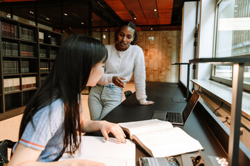 A female student stands and talks to a female student sitting next to her at a table and looking at a book in front of her