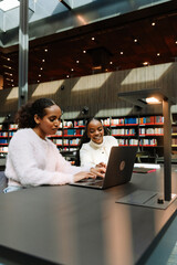 Two female students are sitting at a table and looking at a laptop, one of them typing while the other laughs
