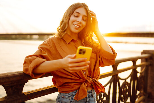 A smiling woman with a phone stands on a sunny embankment. A beautiful woman looks at her smartphone and enjoys the sunset in the fresh air by the water. Urban concept, technology. Lifestyle. - Powered by Adobe
