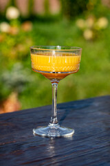 Bright orange cocktail in vintage crystal glass against summer garden background on wooden table, closeup. Delicious orange alcoholic drink for relaxation on a sunny warm day