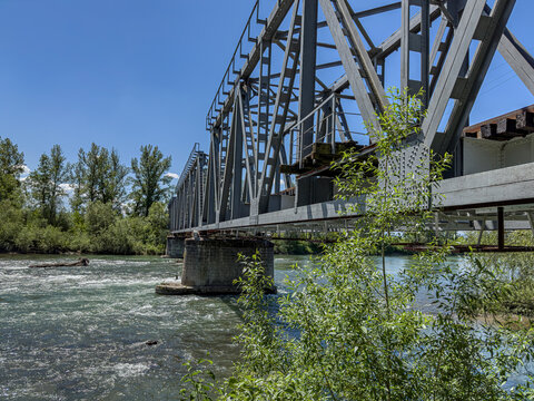 A steel truss bridge spans across a flowing river surrounded by lush greenery under a bright summer sky. The Tisza River. Transcarpathia, Ukraine. - Powered by Adobe