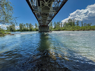 A steel truss bridge stretches over a flowing river with clear reflections and surrounding trees under a bright sky with clouds. The Tisza River. Transcarpathia, Ukraine.
