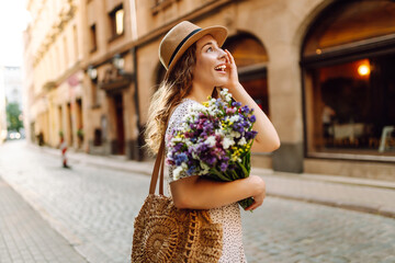 Cheerful woman in hat strolls along sunny street with bouquet of wildflowers. Beautiful woman holds bouquet and enjoys city atmosphere. Conceptual concepts for walking, recreation, and tourism.