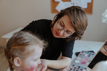 A young mother and her 3-year-old daughter play dress-up as makeup artists, sharing laughter and...