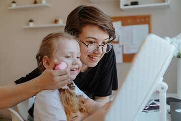 A young mother and her 3-year-old daughter play dress-up as makeup artists, sharing laughter and...