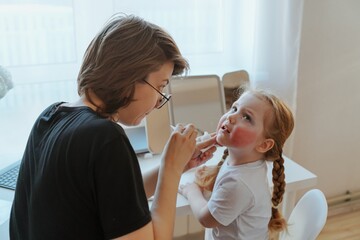 A young mother and her 3-year-old daughter play dress-up as makeup artists, sharing laughter and...