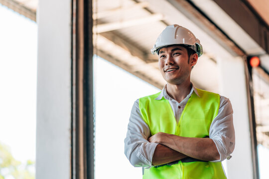 A male warehouse manager in a white shirt, a white hard hat, and a neon green reflective vest stands with his arms crossed. He is looking camera.a sense of professional supervision and order.
