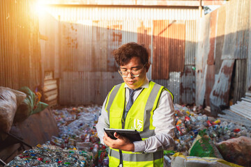Professional waste management worker using tablet device in recycling facility surrounded by plastic bottles and containers in urban environment