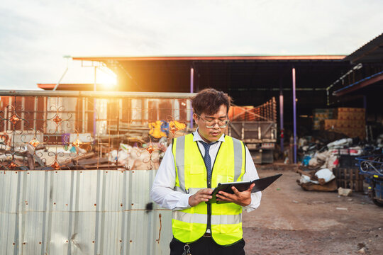 Professional Asian man in safety vest checks tablet device at junkyard during sunset, overseeing recycling and waste management operations in urban area - Powered by Adobe