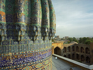 Aerial view of intricate blue and turquoise tilework adorning a historic building with Arabic script, contrasting against the soft, blurred cityscape, Samarkand, Uzbekistan.