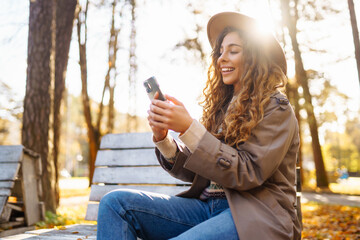 Portrait of cheerful woman in hat, using phone, sitting on bench in autumn park. Beautiful woman enjoys sunny day, using phone on wooden bench. Concept of blogging, relaxation, and seasonality.