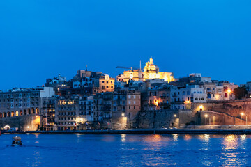 Obraz premium Panoramic twilight view of Valletta’s historic waterfront with illuminated bastions, limestone buildings, and a church dome glowing above the Mediterranean Sea