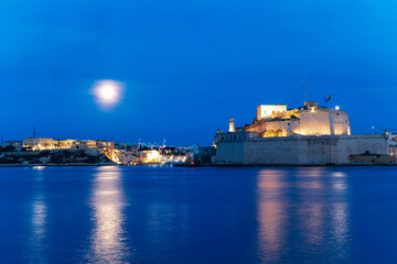 Fototapeta premium Illuminated Fort St. Angelo stands on the Grand Harbour at twilight in Malta, its limestone bastions glowing beneath a blue evening sky and full moon, with reflections shimmering on the water.