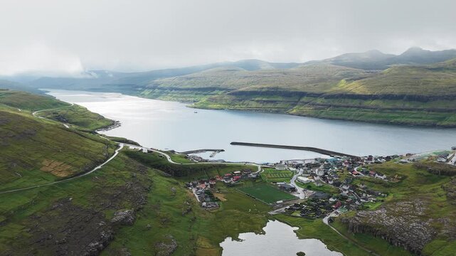 Aerial view of Ei&eth;i, a charming village nestled amidst green hills and a serene blue fjord, revealing a picturesque harbor and scattered houses, Ei&eth;i, Eysturoy, Faroe Islands.