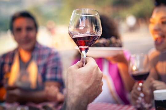 Close up of a man holding a glass of red wine during a toast at a party. Concept of winetasting, celebration and Italian lifestyle with people laughing in the background.