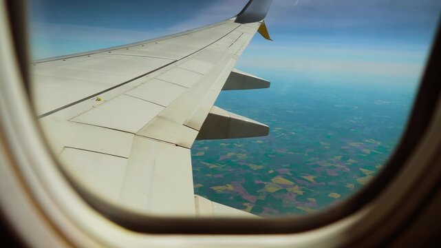 Jet flying above patchwork farmland with scattered fields, view from window porthole. Plane cruising over colorful agricultural landscape with divided plots. Aircraft gliding above countryside with