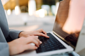 Close-up of a businesswoman typing on a laptop against the backdrop of a modern business center. A young woman working on a laptop outdoors. Concept of modernity and technology. Cozy atmosphere.