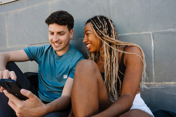 A female athlete looks at a male athlete who is holding a phone while they laugh and lean on a wall
