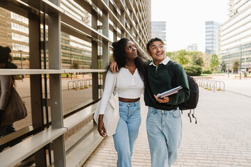 Male student holding notebooks and hugging female student walking next to him while they laugh and...