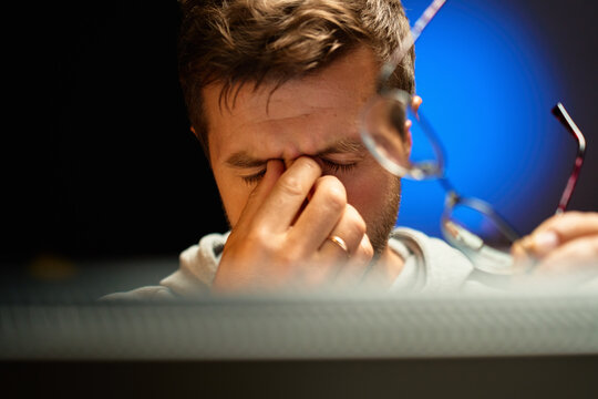 Close-up of tired man rubbing his eyes and holding glasses while sitting at computer. Person has fatigue and headache from overwork and eye strain