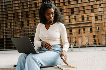 A female student sits and opens a book while listening with earphones and a laptop on her lap