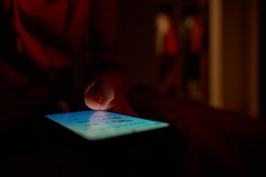 Man using smartphone at night. Close up of hand holding smartphone, swiping and scrolling social media feed on glowing screen in dark room