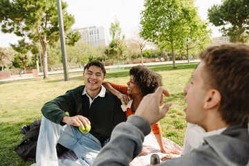 A group of three students sit on a blanket and laugh while two of them hold an apple and a sandwich