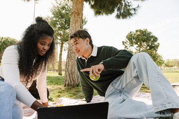 A male student is holding an apple and talking to a female student who is sitting next to him on a blanket and looking at a laptop