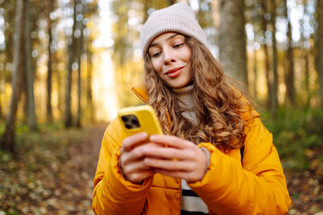 A young woman in a bright jacket with a phone in an autumn forest. A beautiful woman enjoys the sunny weather and nature with her phone outdoors. Concept of nature, technology.