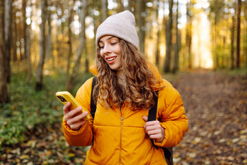 A young woman in a bright jacket with a phone in an autumn forest. A beautiful woman enjoys the sunny weather and nature with her phone outdoors. Concept of nature, technology.