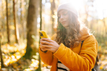 A young woman in a bright jacket with a phone in an autumn forest. A beautiful woman enjoys the sunny weather and nature with her phone outdoors. Concept of nature, technology.