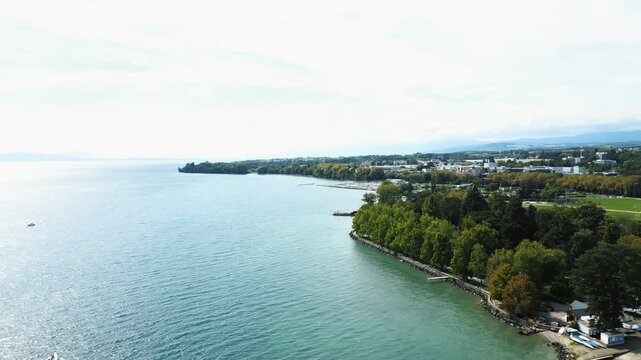 Aerial view of the shoreline, where turquoise waters meet lush green trees and buildings, creating a vibrant contrast of colors, Lake Geneva, Morges, Vaud, Switzerland.