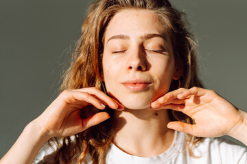 Obraz premium A portrait of a sweet woman touching her face in the sun's rays. A beautiful woman enjoys natural beauty in a cozy room. Concepts of beauty, grooming, and naturalness.