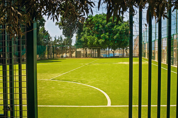 A mini football field with artificial turf sits behind a fence in a park. Lush trees, the sea, and a clear sky are visible in the background.