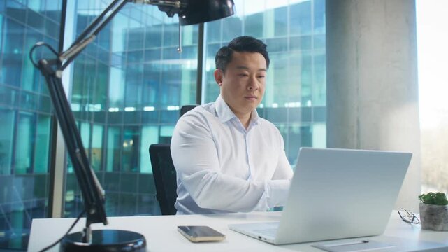 Focused Asian man in elegant shirt putting on headphones while opening laptop in daylight office. Getting ready for virtual meeting or online call. Preparing to join conference or start webinar.